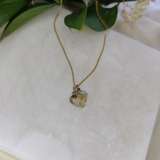 Gold necklace with a pendant on a white marble surface with green leaves and flowers.