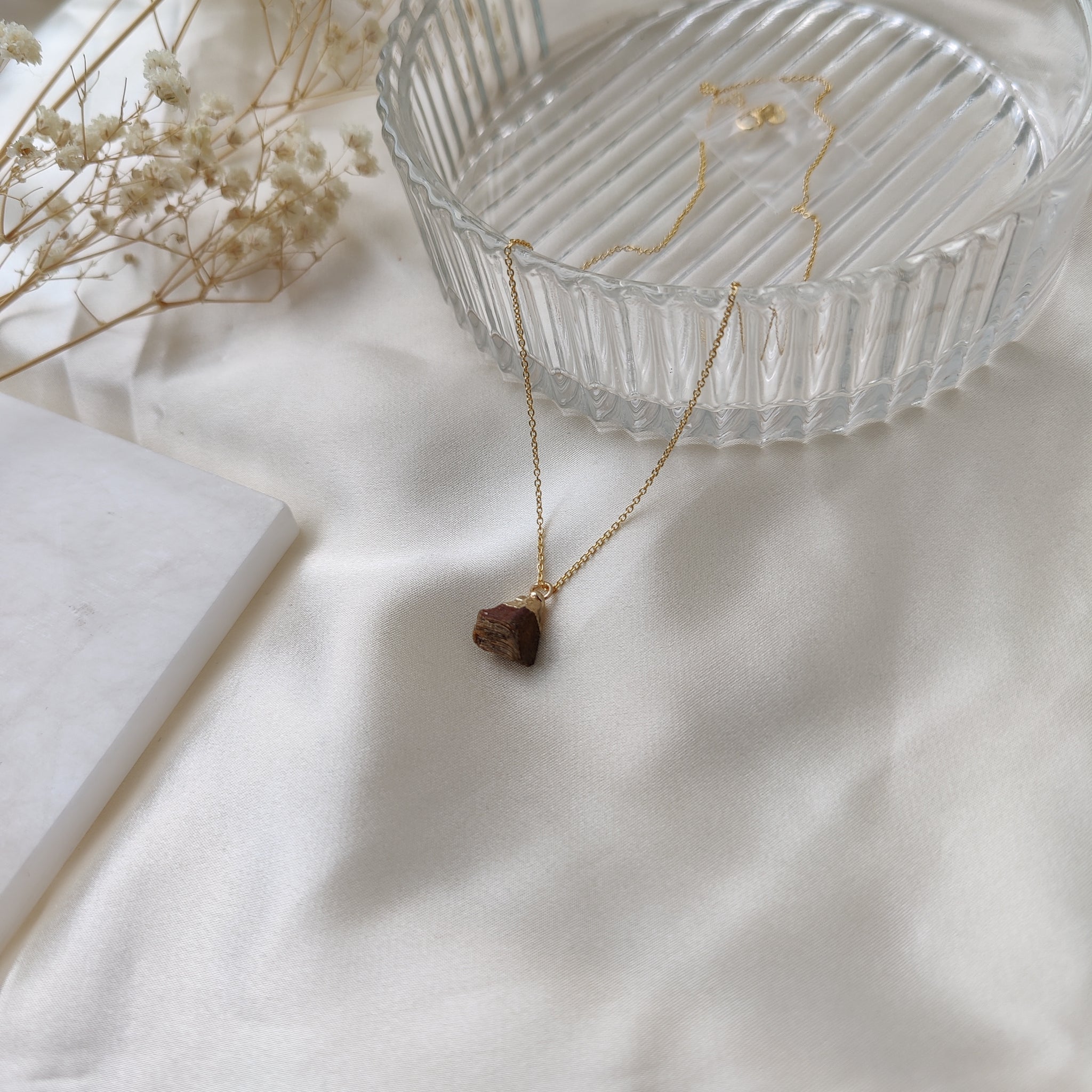 Necklace with a pendant on a white surface with a glass dish and dried flowers.